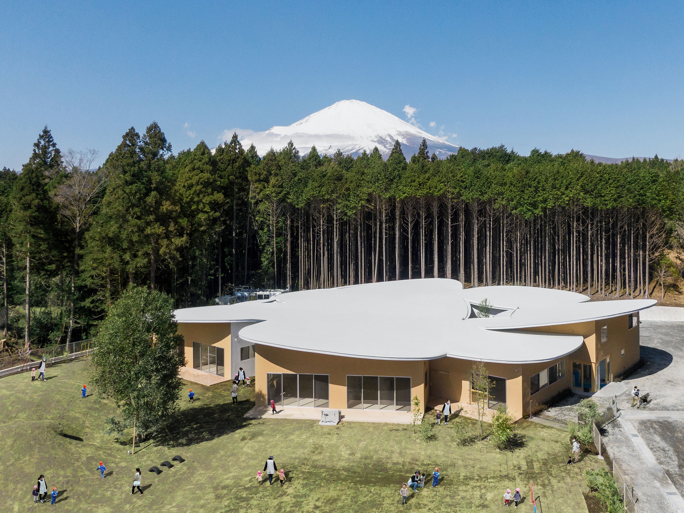 山下貴成建築設計事務所による、静岡・御殿場市の「高嶺の森のこども園」。森の中の起伏に富む地形の敷地。各年齢の子供達の生活が“同時に満たされる”在り方を求め、周辺環境に応じて８つの部屋を“円環状”に配置する構成を考案。地形と呼応し“湾曲する”屋根で全体を緩やか  ...